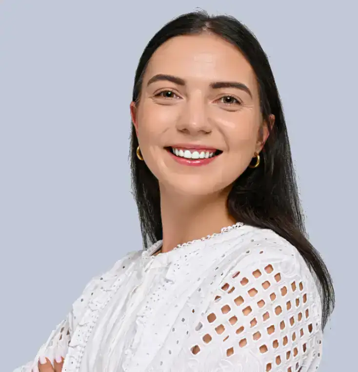 A woman with long dark hair, wearing a white textured blouse with cut-out details and gold hoop earrings, smiles confidently against a light gray background.