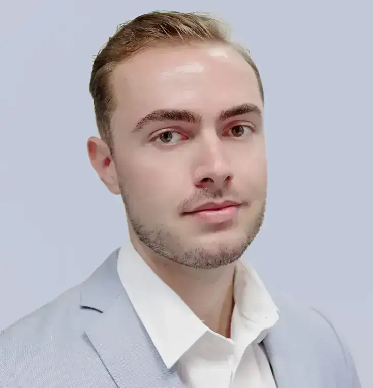 A young man with short light brown hair and trimmed beard, wearing a light blue blazer and white shirt, poses against a plain light blue background.