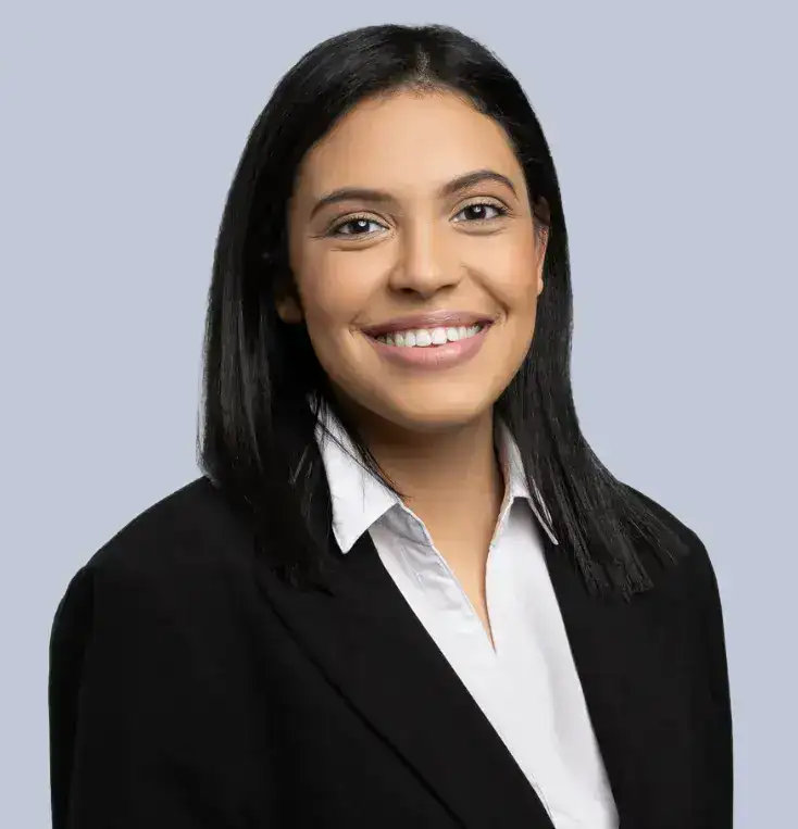 A woman with straight, shoulder-length black hair smiles at the camera. She is wearing a white collared shirt and a black blazer, with a light gray background behind her.