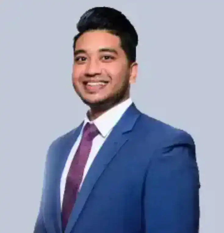 A man with short dark hair, wearing a blue suit, white shirt, and purple tie, smiles while standing against a plain light grey background.