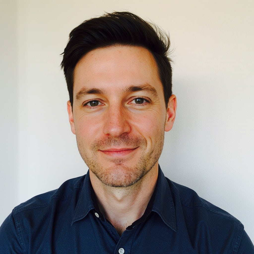A man with short, dark hair and light skin smiles slightly at the camera. He is wearing a dark blue, button-up shirt and is posed in front of a plain, light-colored background.