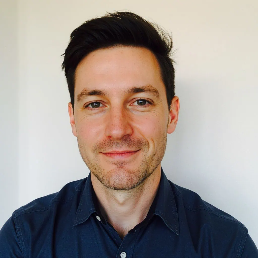 A man with short, dark hair and light skin smiles slightly at the camera. He is wearing a dark blue, button-up shirt and is posed in front of a plain, light-colored background.
