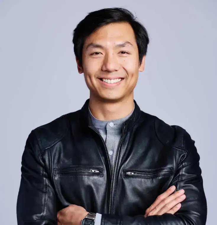 A man with short black hair wearing a black leather jacket and a gray shirt stands with arms crossed, smiling at the camera against a plain light gray background.