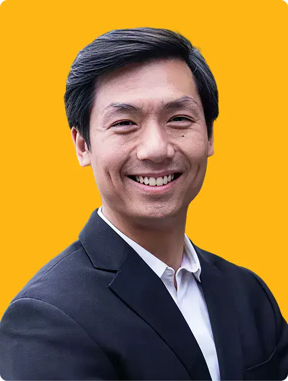 A man with short black hair wearing a black suit jacket and white shirt smiles at the camera against a solid yellow background.