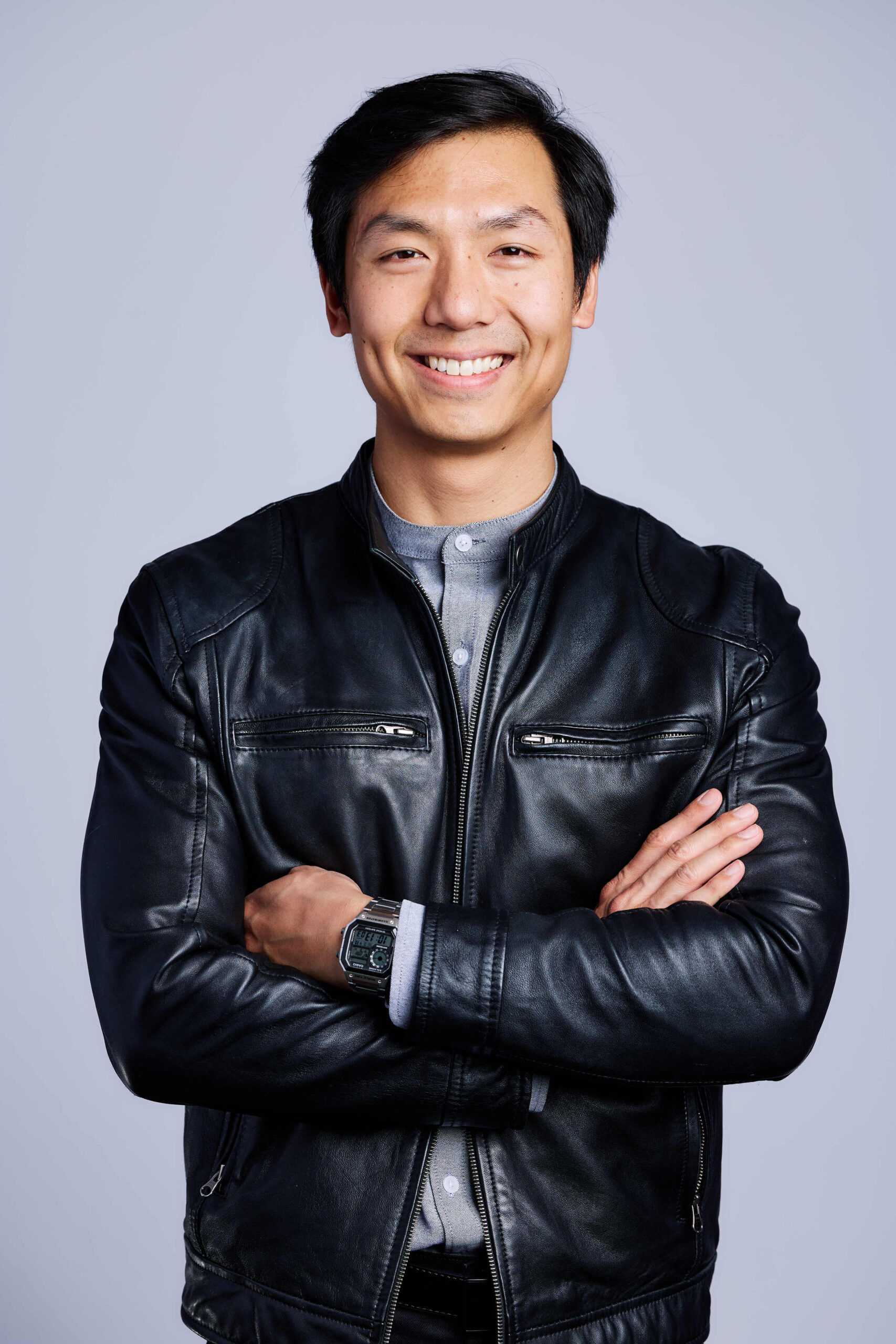 A man with short black hair smiles confidently with arms crossed, wearing a black leather jacket and a wristwatch, standing against a plain light gray background—reflecting the assurance you expect from top SOC Auditors.