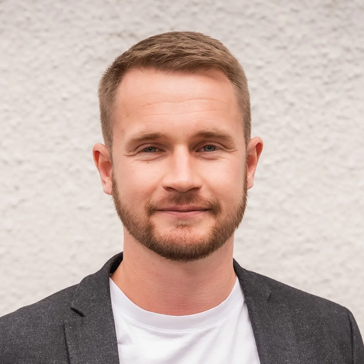 A man with short light brown hair and a beard is wearing a white shirt and a dark blazer, standing in front of a light-colored, textured wall. He is looking at the camera with a slight smile.