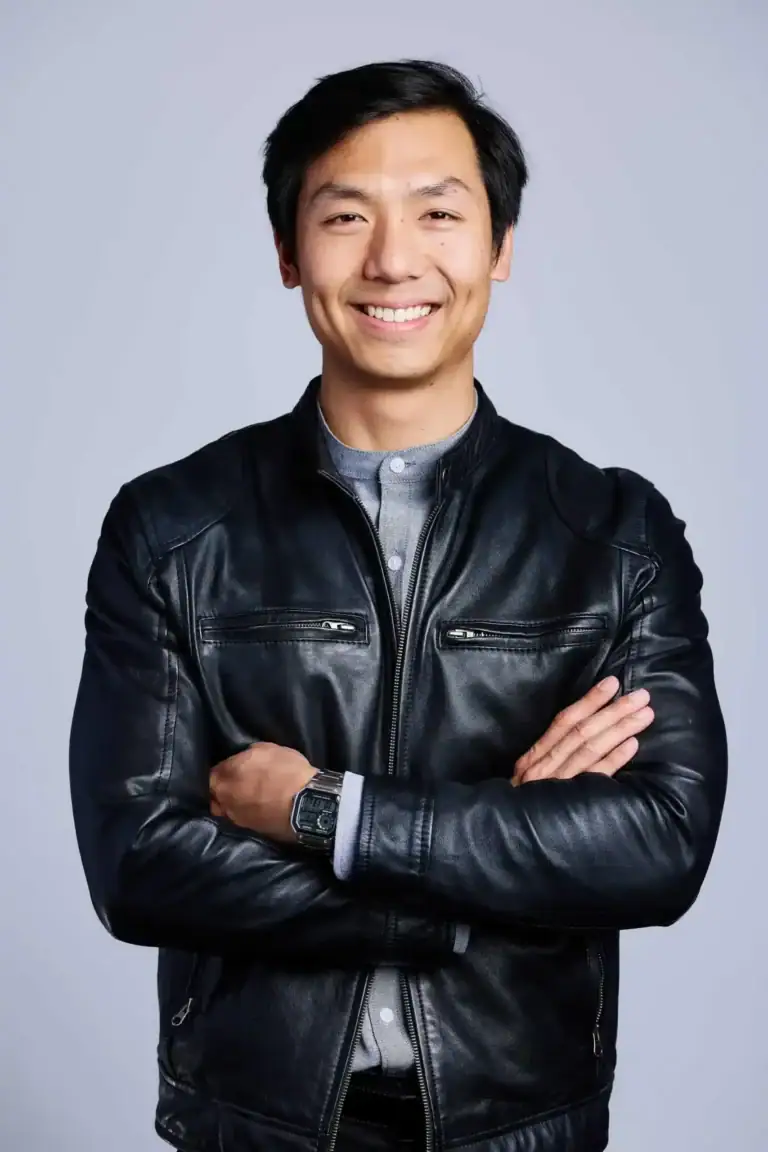 A man with short black hair smiles confidently, standing with arms crossed. He wears a black leather jacket over a gray shirt and a wristwatch, posing in front of a plain light gray background—exuding the professionalism and assurance you’d expect from leading SOC Auditors or an expert at a top SOC 2 audit firm.